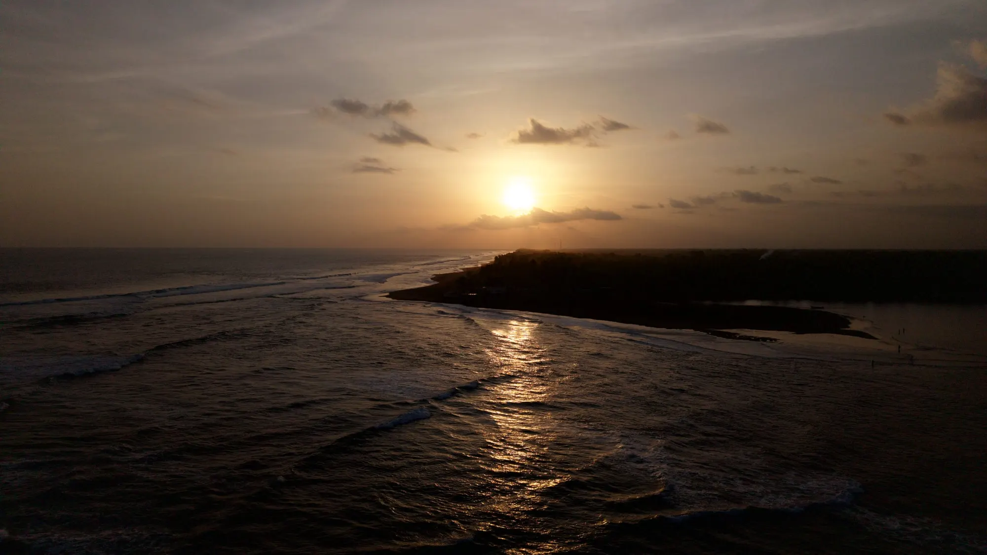 An aerial view at sunset along the Barra de Santiago spit, with lines of Pacific swell breaking on the left and the still estuary water behind the wooded sandbar on the right