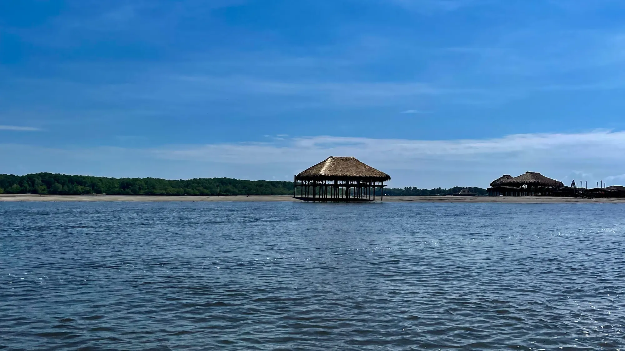 Two large thatched palapas standing on stilts on a sandbar in the estuary, with a mangrove treeline behind and open blue sky above
